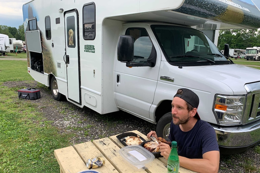 Lenihan sitting at outside table eating food with RV and caravan park in background.