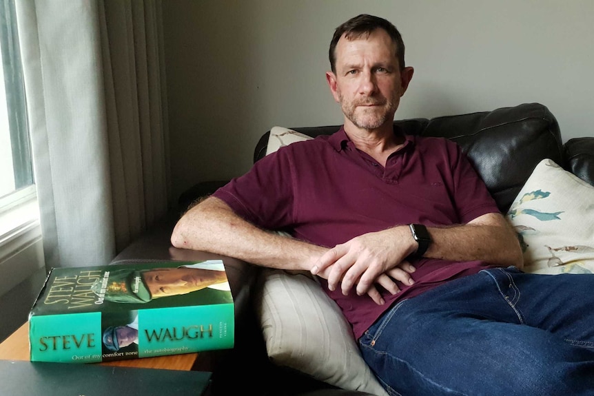 A middle-aged man sitting on a leather couch. To his right, a copy of Steve Waugh's autobiography sits on the table.