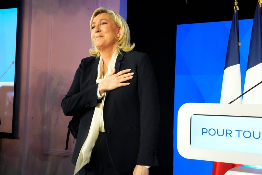 A blonde woman in dark suit places her right hand over her heart as she gestures after speaking at a podium.