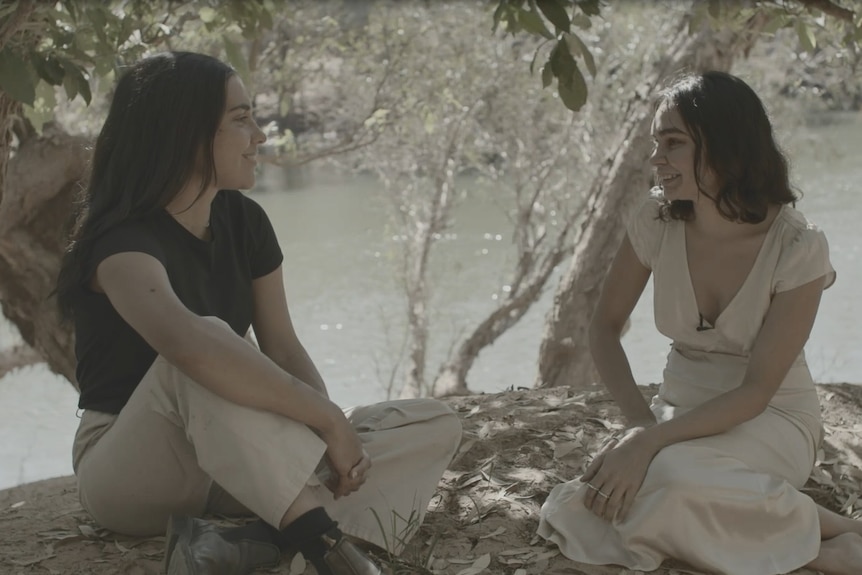 Two young women sit together on the banks of a river. 