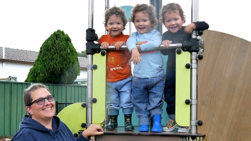 Identical triplet boys smiling at the camera standing next to each other on playground equipment. A woman standing near them