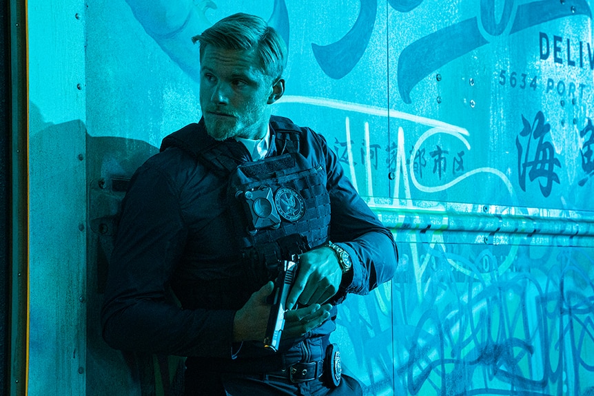 A man wearing bulletproof vest and police attire stands holding pistol against graffitied wall illuminated by light blue light.