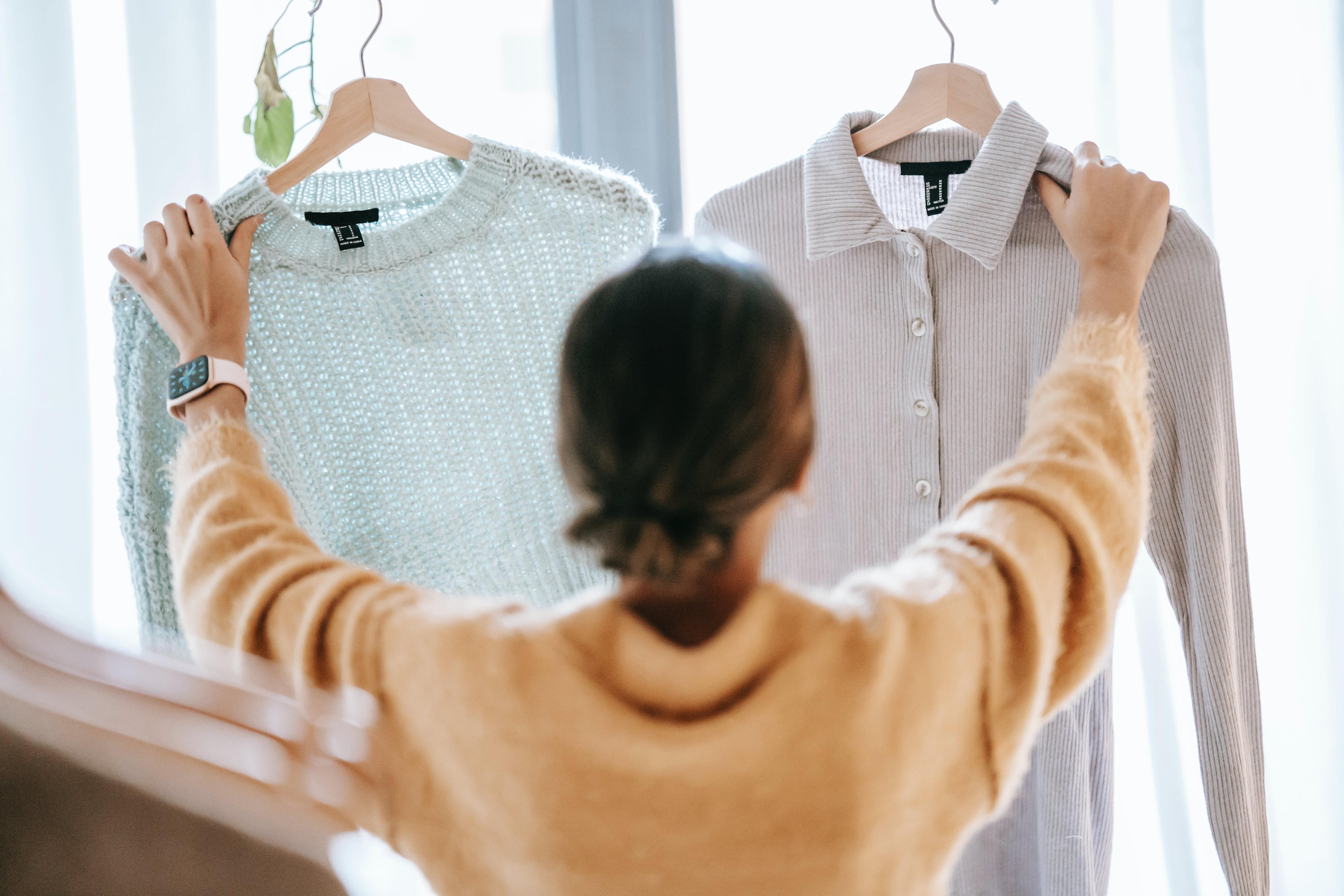 A woman wearing an ochre jumper compares a collared cloth shirt with a blue knitted jumper. The photo is taken from behind.