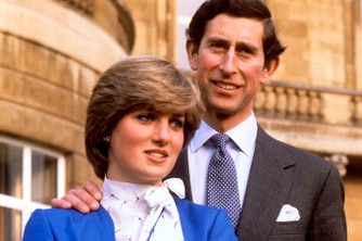 Lady Diana in a blue jacket and skirt with Prince Charles standing behind her, hand on shoulder.