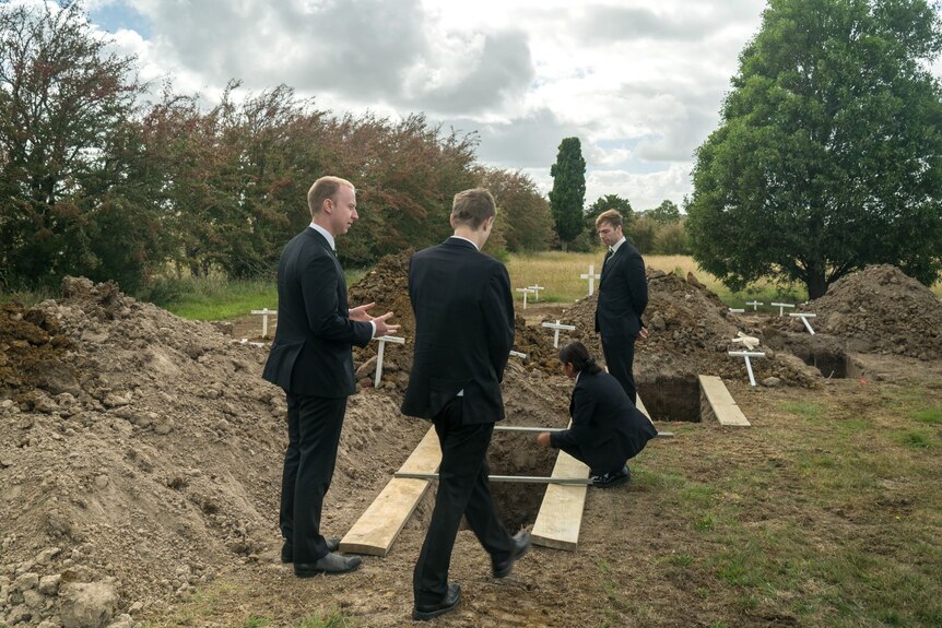 Four people in black suits stand around a recently dug grave which is yet to be filled in with dirt