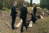 Four people in black suits stand around a recently dug grave which is yet to be filled in with dirt
