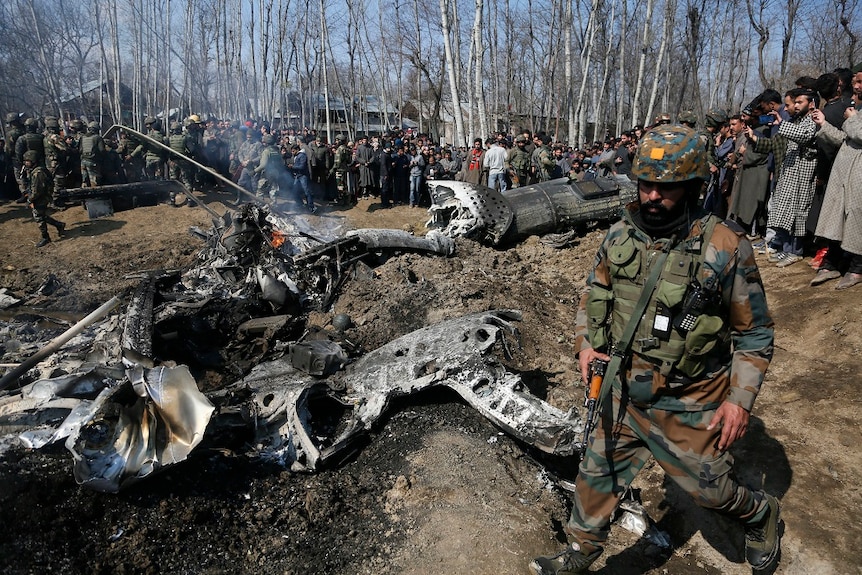 An armed soldier walks past the smoking wreckage of an aircraft as a large crown gathers around
