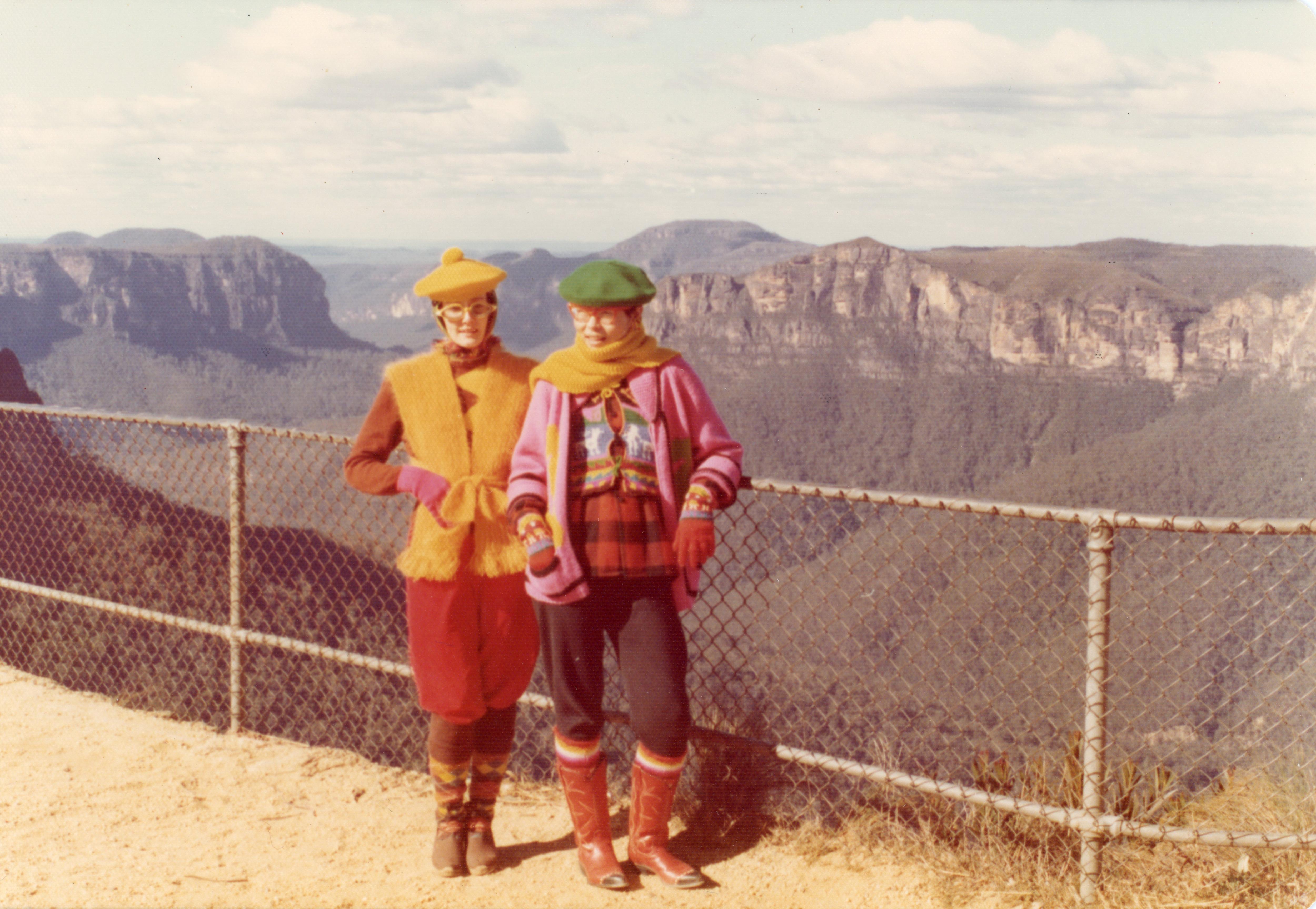 Two women wearing bright jumpers scarves and hats at a mountain range lookout.