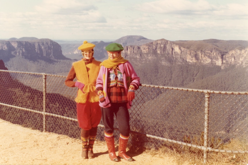 Two women wearing bright jumpers scarves and hats at a mountain range lookout.