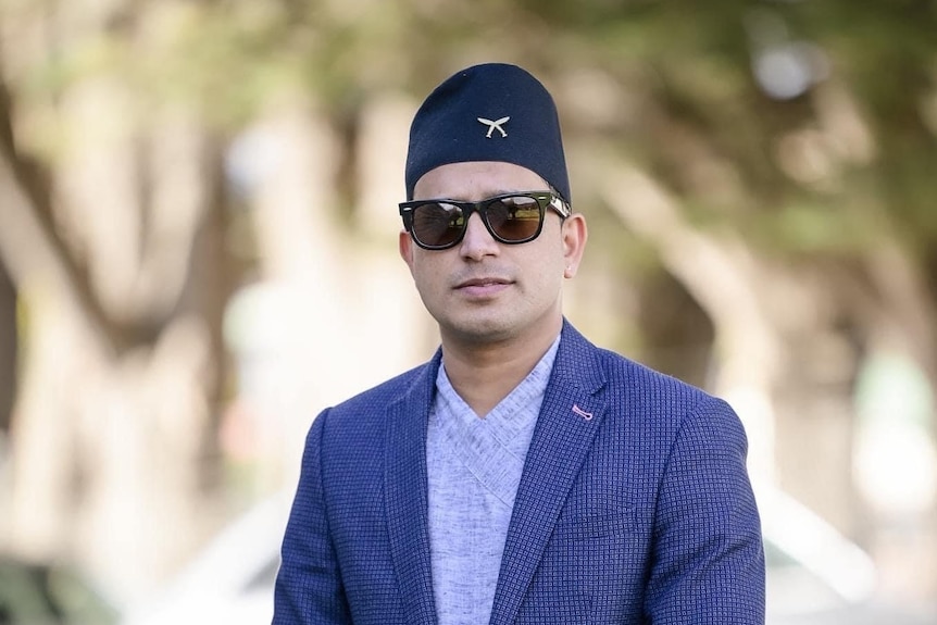 A man stands in semi-traditional Nepali clothing.  He wears a blue hat, jacket and top.