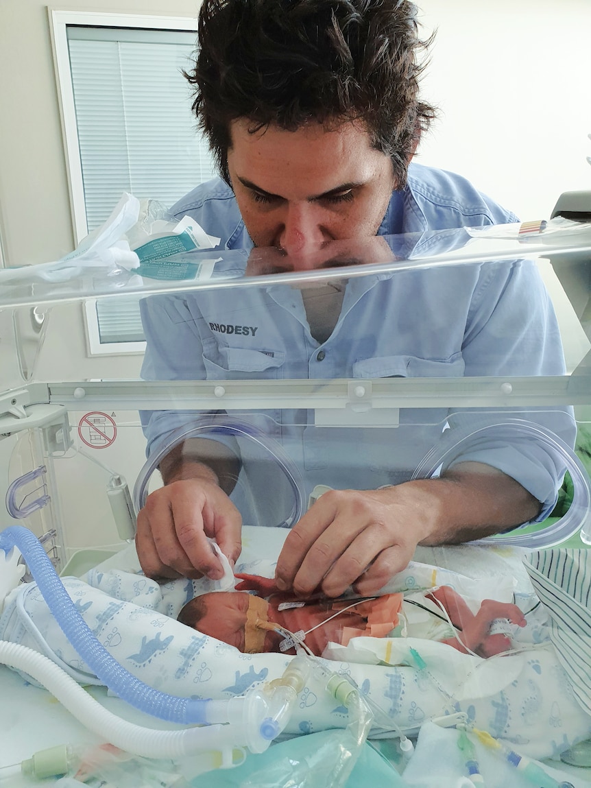 A man stands over a premature baby in its bassinet, washing his baby.