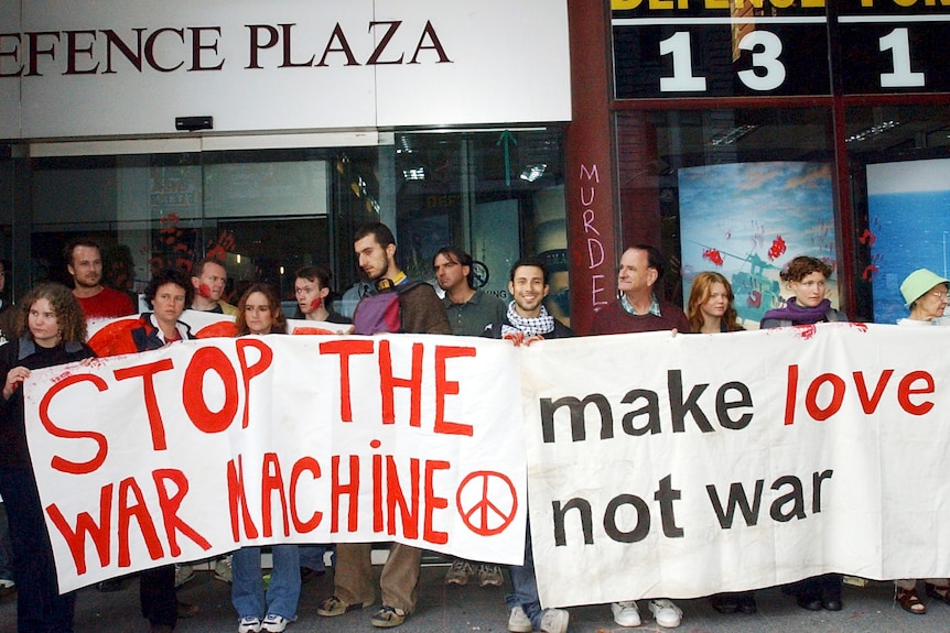 A group hold large banners in front of a building called Defence Plaza. One reads "Stop the war machine" in red paint