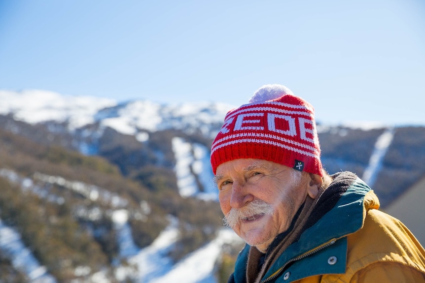 A close up photo of an elderly man wearing a red beanie smiling on a snowy mountain