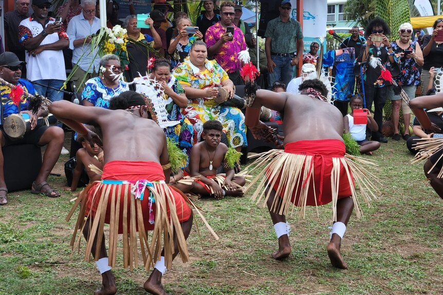 Two First Nations men crouch low as they dance in straw skirts to a crowd watching them.