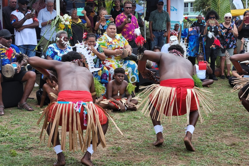 Two First Nations men crouch low as they dance in straw skirts to a crowd watching them.