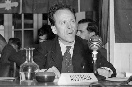 A black and white image of a man in a suit sitting behind a microphone. 