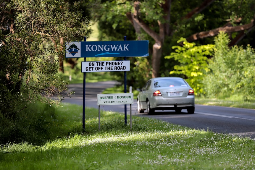 Signs reading 'Kongwak' and 'On the phone Get off the road' on a lush green road side as a sedan passes while braking