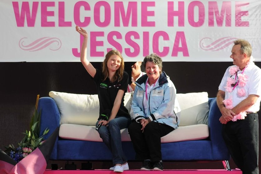 Jessica and her parents sitting on a couch, Jessica waving, wall behind says "Welcome home Jessica".
