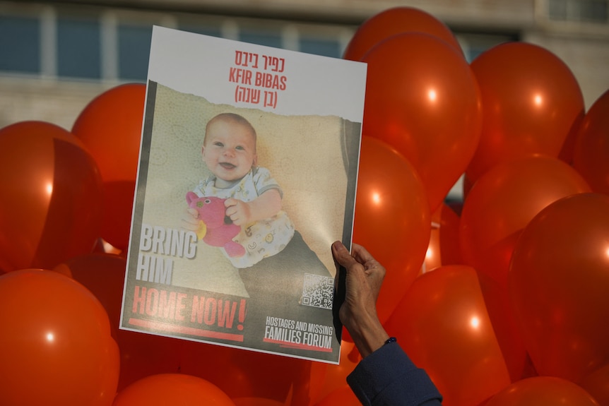 A close up of balloons, in front of which a hand holds up a poster which shows a baby boy and the words 'bring him home now'