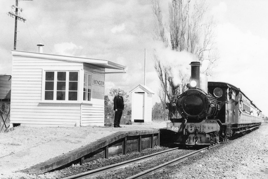 A black and white photo of a steam locomotive at an old station with a rail guard standing alongside. 