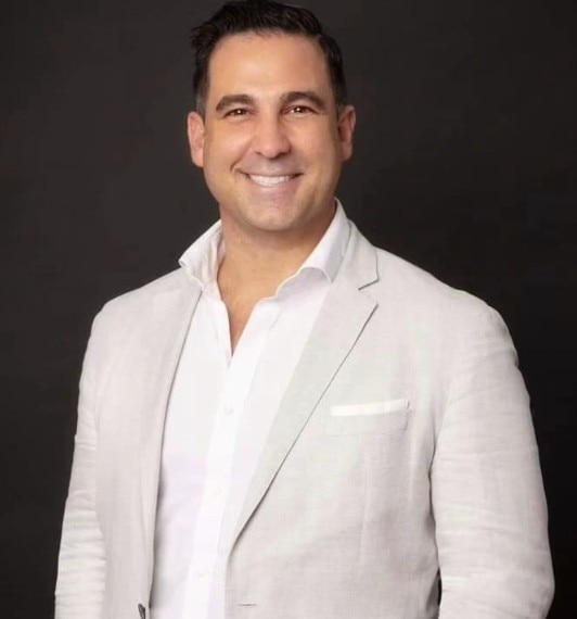 A headshot of a smiling man wearing a white shirt and light grey jacket.