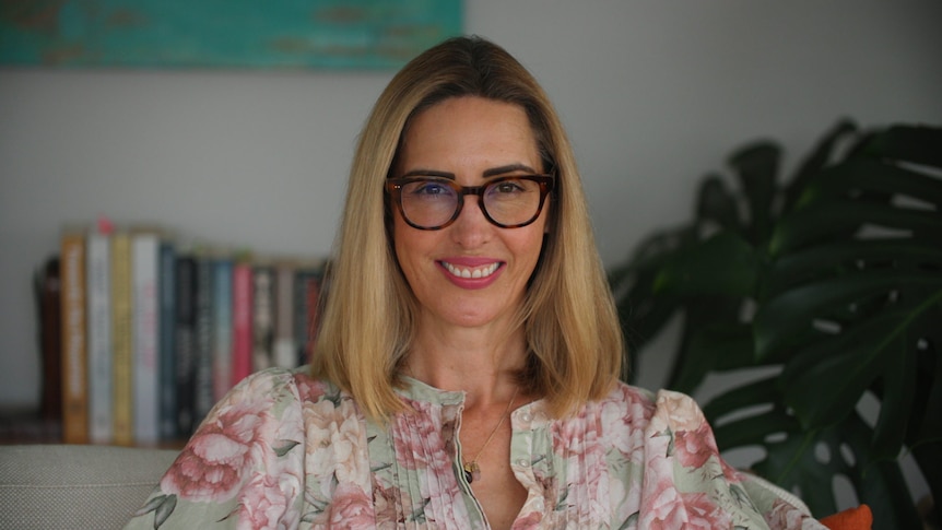 A woman in a floarl blouse and glasses looks at the camera