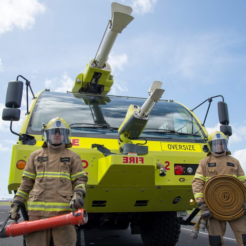 Two aviation firefighters stand in front of a specially designed, bright yellow fire truck.