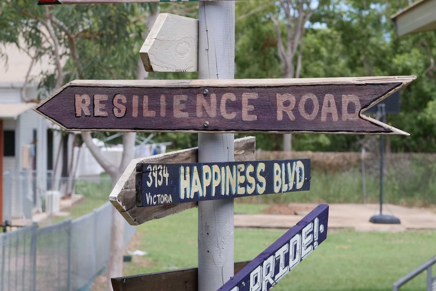 Wooden signs reading 'resilience road', 'happiness road' and 'pride' pointing in different directions and nailed to a post.