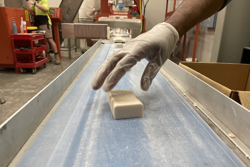 A worker's hand is seen placing a bar of soap on a blue conveyor belt inside a recycling facility.