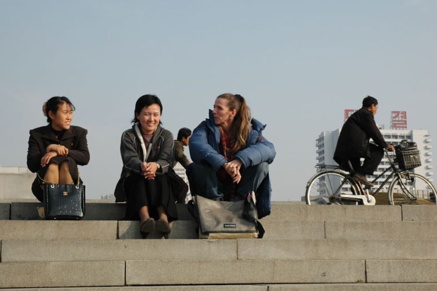 A western woman wearing a blue jacket sits on a step talking to two Asian women as a man rides a bicycle in the background