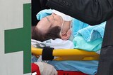 Jae-myung lies on a stretcher with a white face mask and blue blanket on.