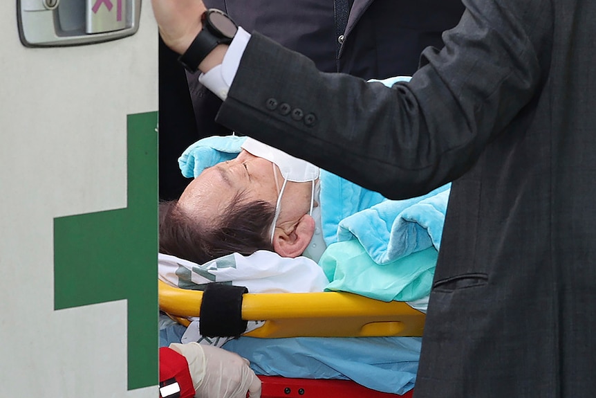 Jae-myung lies on a stretcher with a white face mask and blue blanket on.