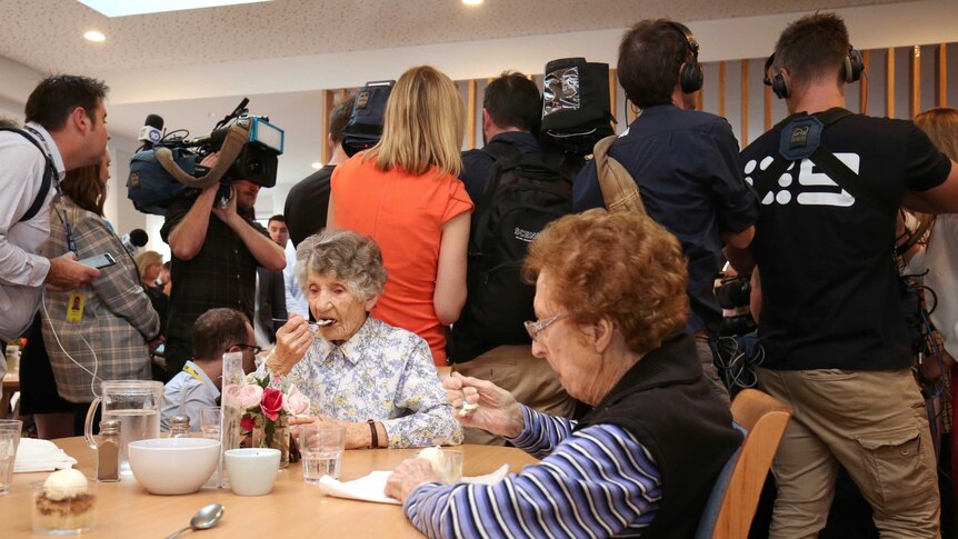 Two women sitting at a table eat their dessert paying no attention to the media pack behind them