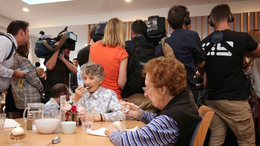 Two women sitting at a table eat their dessert paying no attention to the media pack behind them