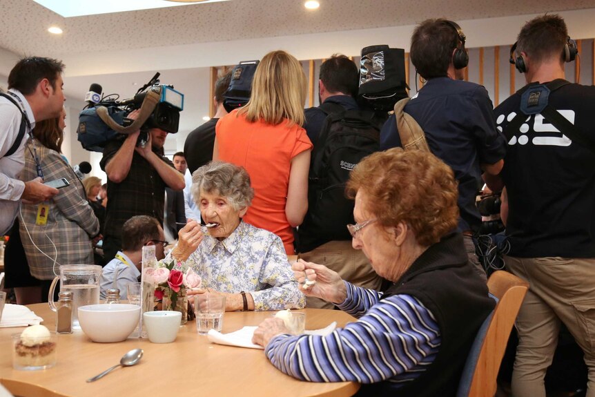 Two women sitting at a table eat their dessert paying no attention to the media pack behind them