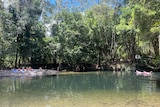 Clear green and blue body of water, with people laying in the sun in the far distance backed onto rainforest setting