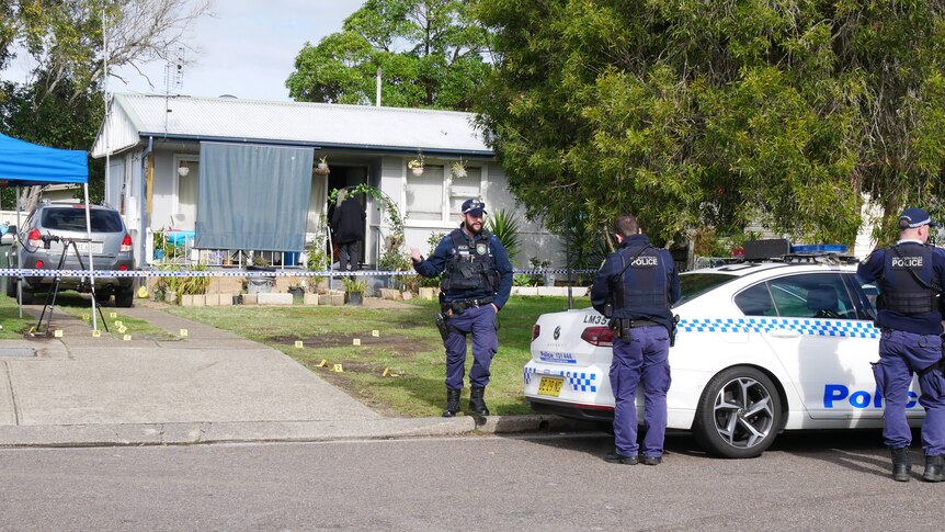 Police man wlaking out the front of a small blue house with police tape and yellow markers 