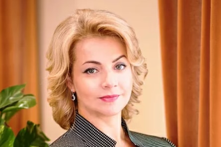 A headshot of Elena Georgieva wearing a black suit jacket with short blonde hair looking at the camera