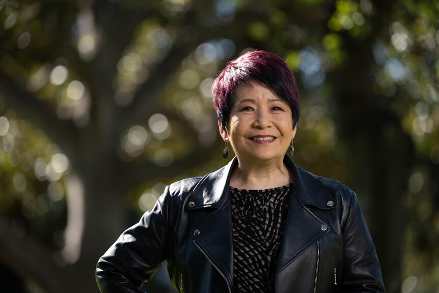 A middle-aged Chinese Australian woman with purple-tinged black hair wears a black leather jacket and smiles in front of trees.