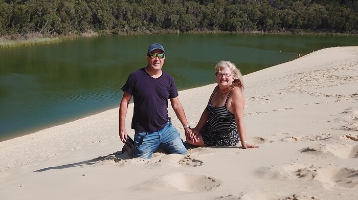 A couple sit on the sand near a lake.