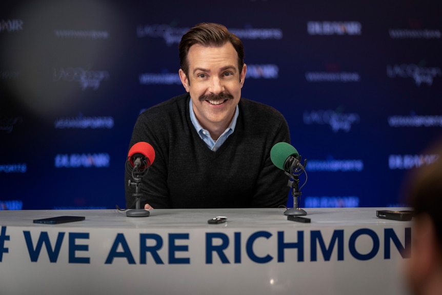 Jason Sudeikis sits at a desk with two microphones in Ted Lasso.