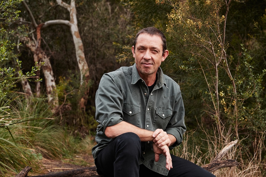 Angus Cerini crouching outdoors with native plants behind him, looking at camera, smiling.