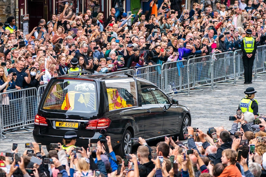 Crowds line both sides of the street as a hearse drives along with a flag-draped coffin in the back