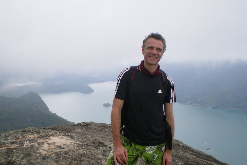 a man in a black adidas top stands on a hill overlooking a river as he looks at the camera and smiles