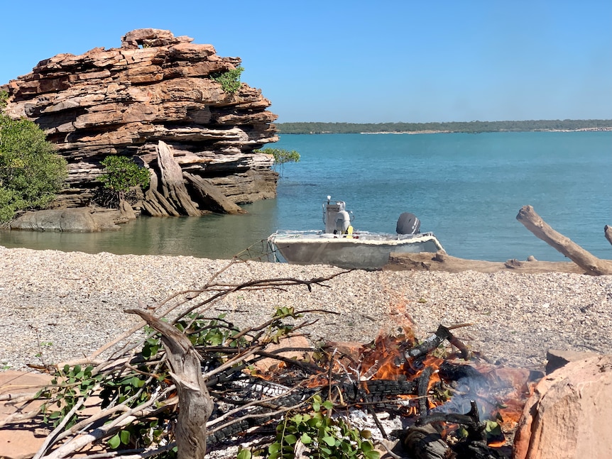 Image of the coast of a remote island, with a campfire in the foreground and boat in the background