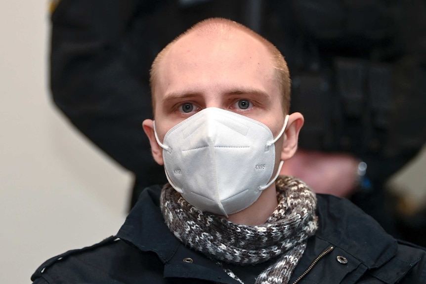 Man with short cropped hair and white face mask stares at camera.