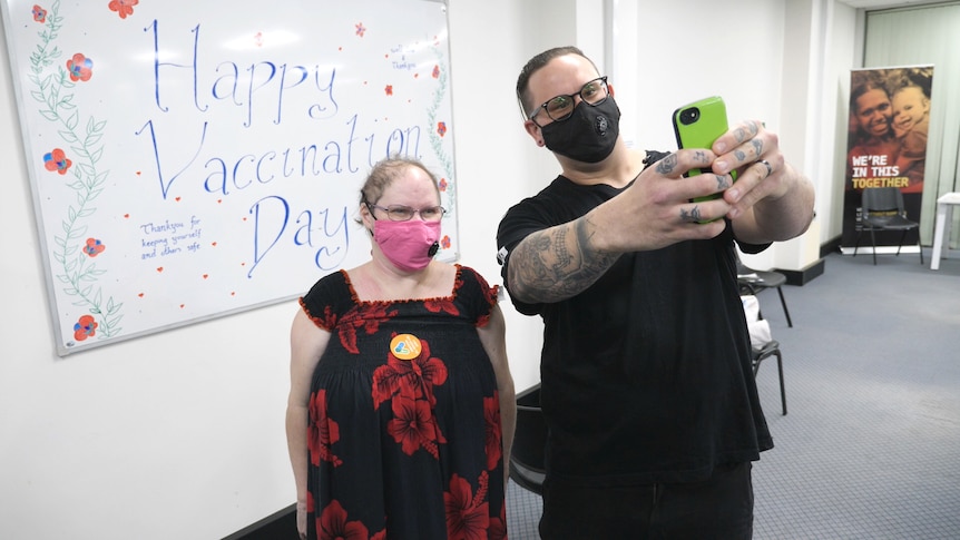 Vaugn taking a selfie of him and Raelene in front of a whiteboard with the words "Happy Vaccination Day", both wearing masks