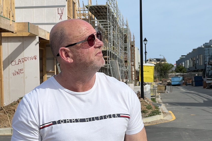 A man wearing a sunglasses looks to the side of the camera. Behind him is a housing development under construction