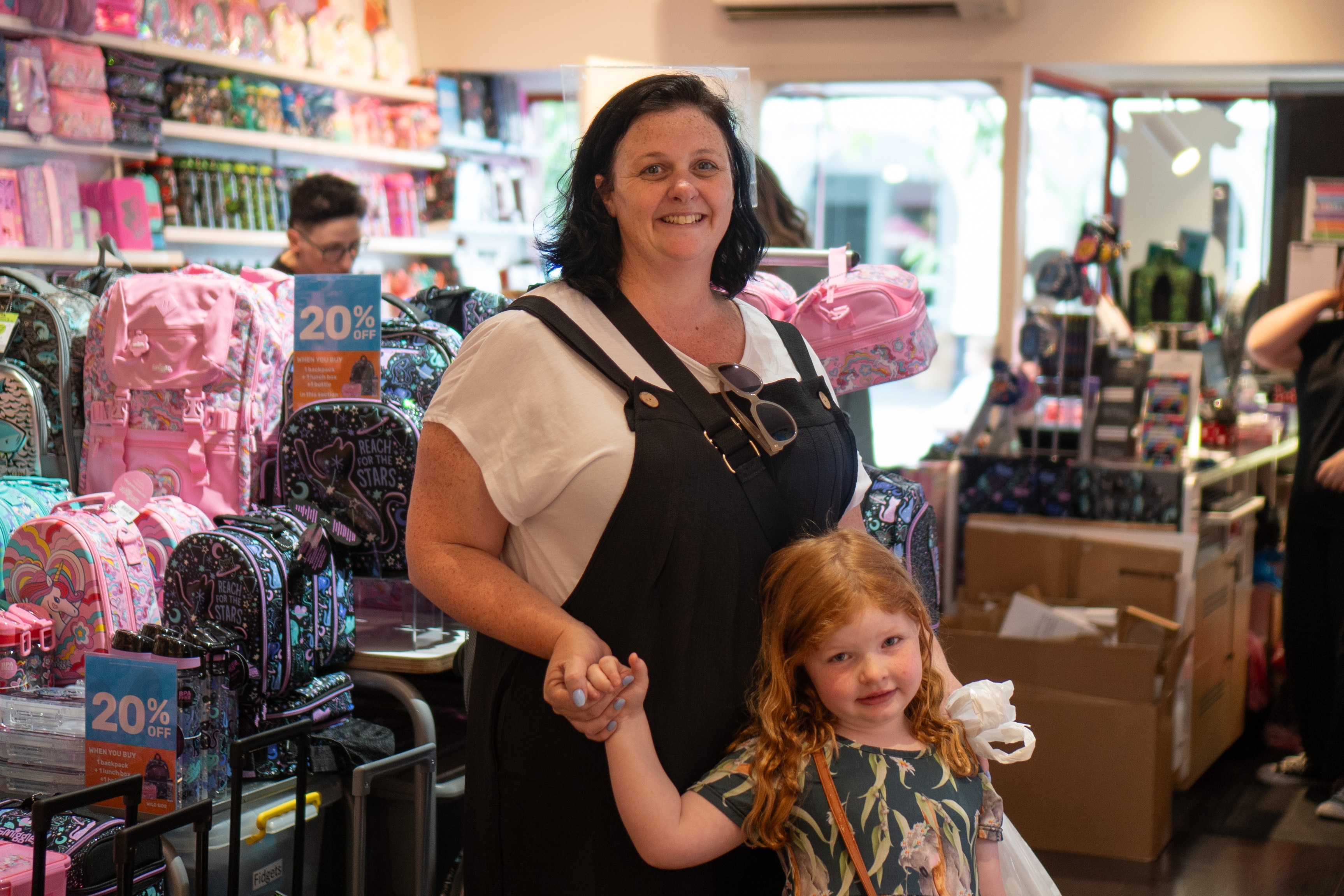 A woman in dungarees and her daughter hold hands in a Smiggle store, smiling at the camera.