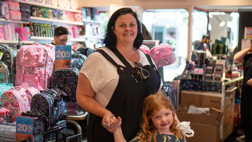 A woman in dungarees and her daughter hold hands in a Smiggle store, smiling at the camera.